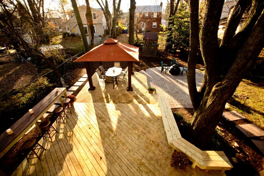 An elevated view of a wooden deck with a bar and stools, overlooking a backyard with a patio, gazebo, outdoor furniture, and leaf-bare trees in sunlight.